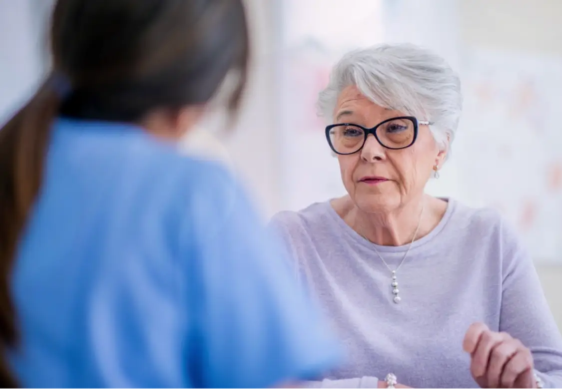 An older woman wearing thick-rimmed glasses and a light purple blouse, talking with a healthcare professional, who has her back turned to the camera. Actor portrayals.