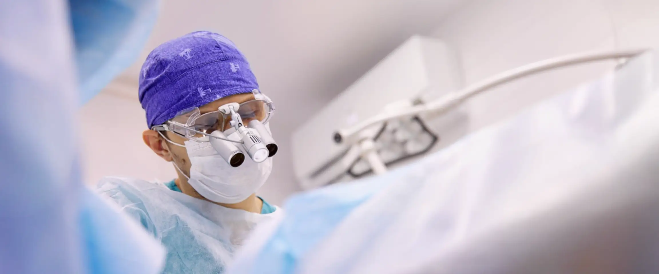 Surgeon wearing full surgical scrubs and wearing magnifying glasses, operating on a patient. Actor portrayal.
