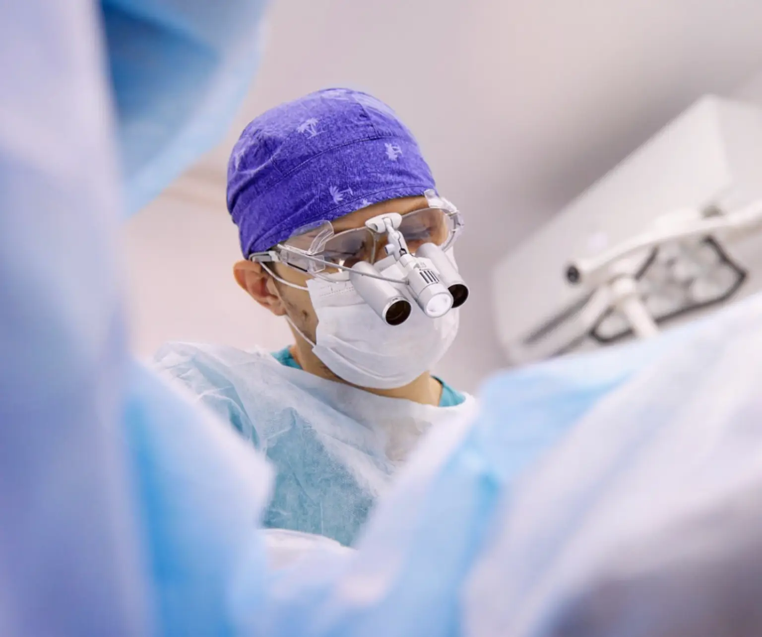 Surgeon wearing full surgical scrubs and wearing magnifying glasses, operating on a patient. Actor portrayal.