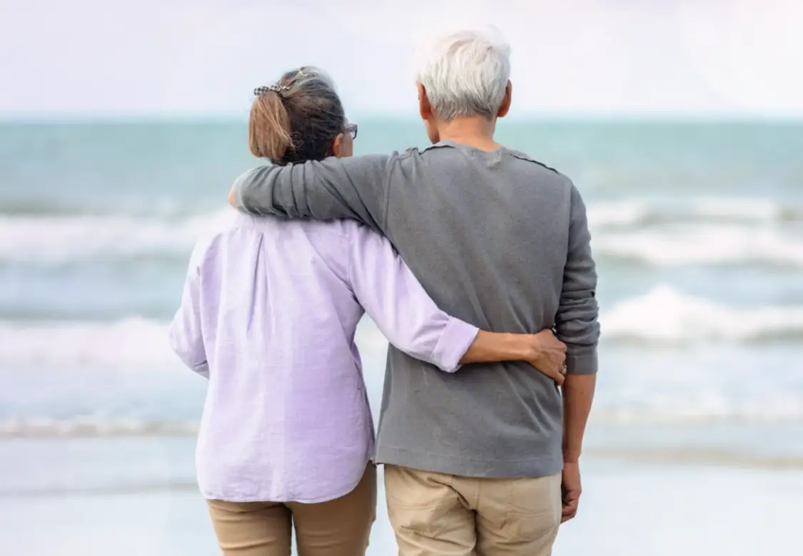 An older man and woman standing arm in arm on the beach. Actor portrayals.