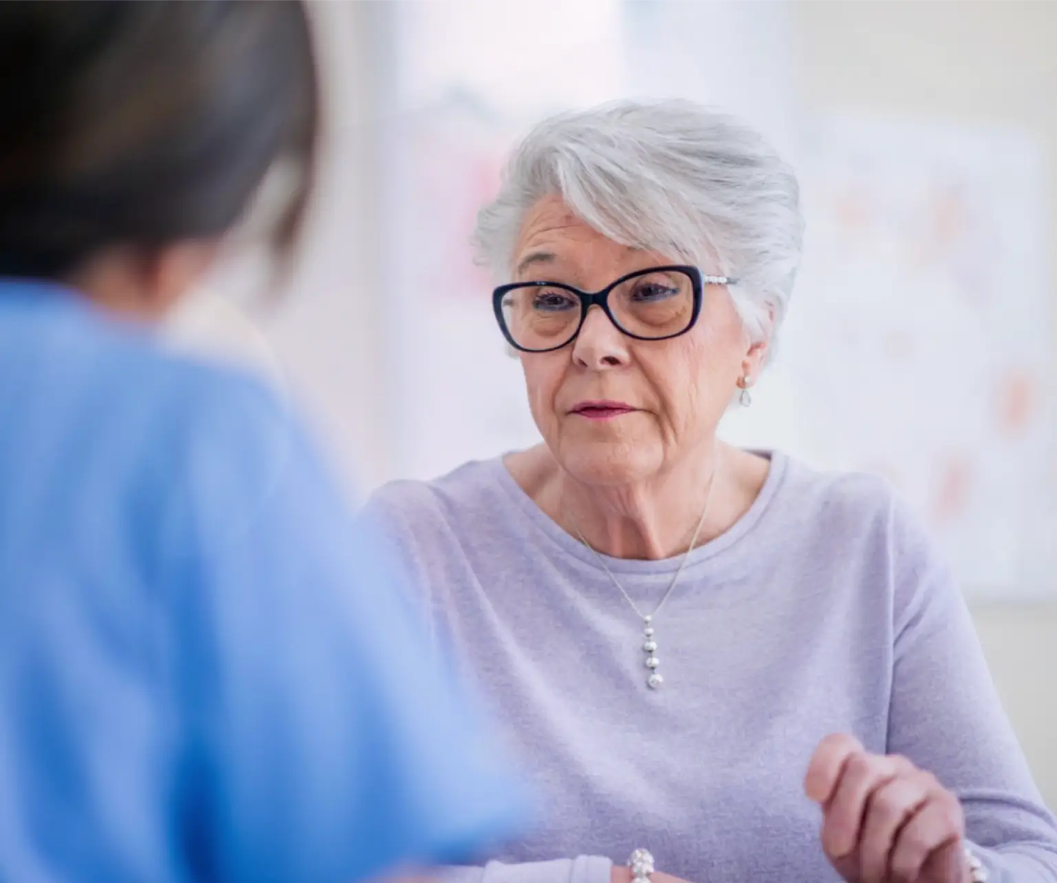 An older woman wearing thick-rimmed glasses and a light purple blouse, talking with a healthcare professional, who has her back turned to the camera. Actor portrayals.