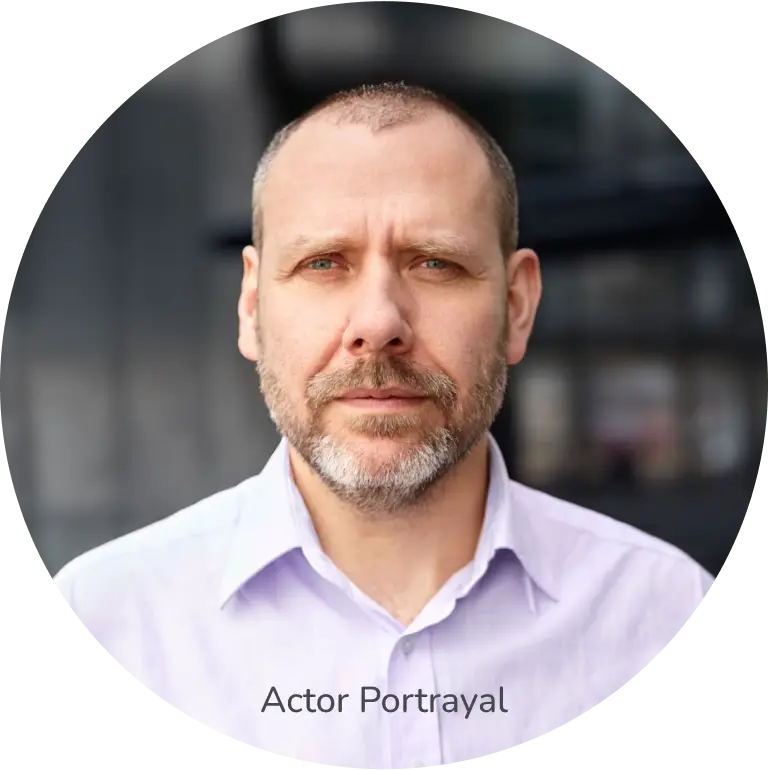 A bearded man, wearing a light purple shirt, looking directly at the camera. Actor portrayal.