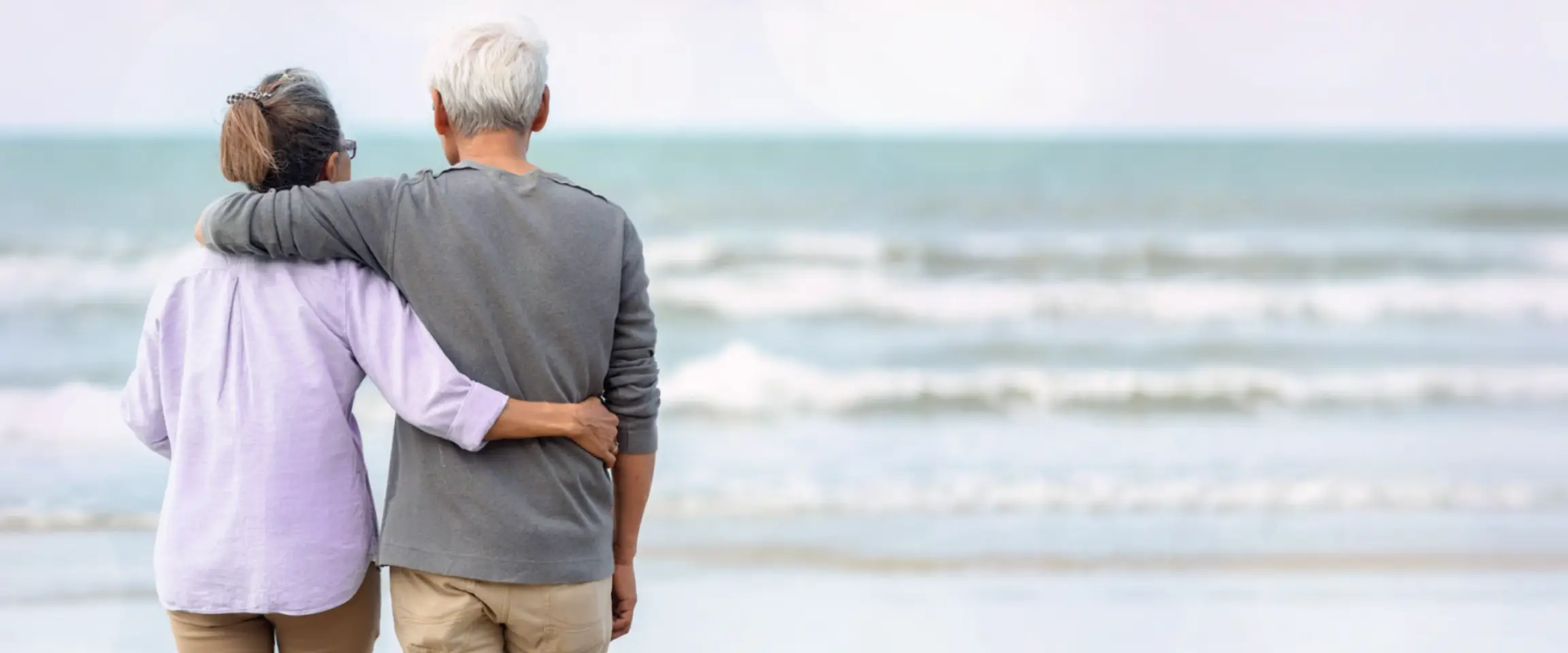 An older man and woman standing arm in arm on the beach. Actor portrayals.