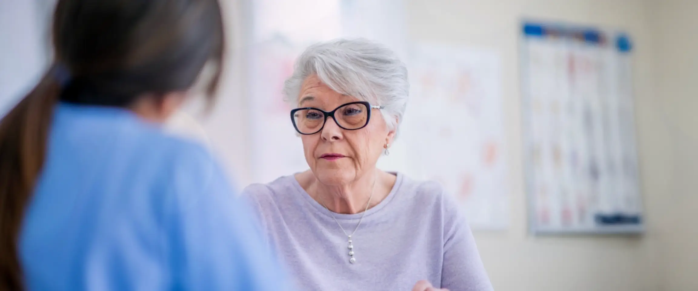 An older woman wearing thick-rimmed glasses and a light purple blouse, talking with a healthcare professional, who has her back turned to the camera. Actor portrayals.