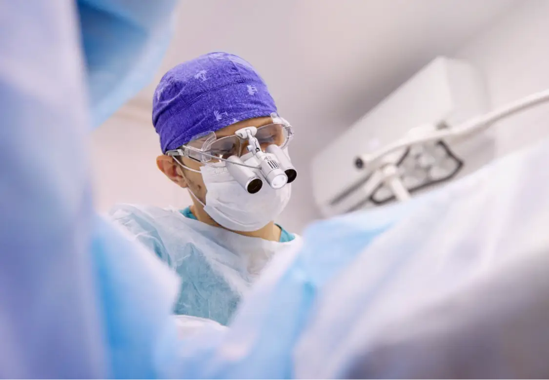 Surgeon wearing full surgical scrubs and wearing magnifying glasses, operating on a patient. Actor portrayal.