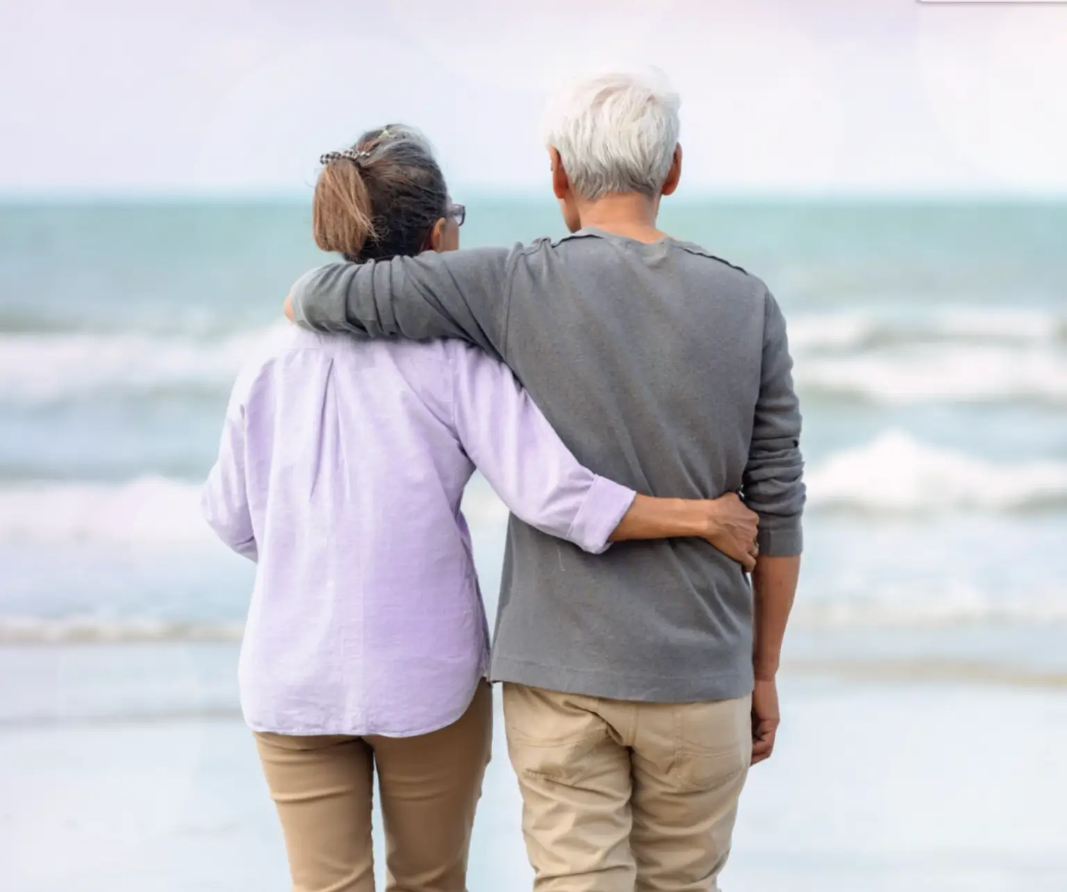 An older man and woman standing arm in arm on the beach. Actor portrayals.